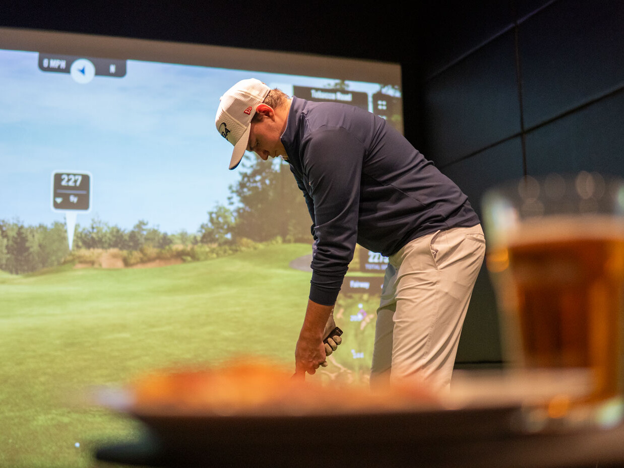 Golfer Lines up Shot in Front of a Indoor Golf Simulator Screen. Food and Drink in the Foreground